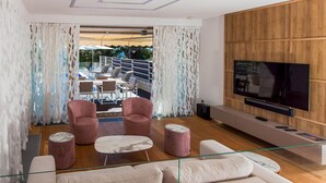 A modern living room with a sofa, two armchairs, a coffee table, and a TV. There is a balcony with outdoor seating visible through sheer curtains. - Beach front home in Petrcane (Petrcane)
