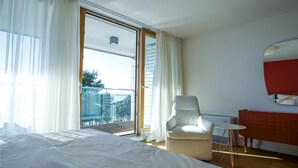 A modern hotel room with a large bed, a white armchair, a small table, and a view of the city through a large window. - Beach front home in Petrcane (Petrcane)