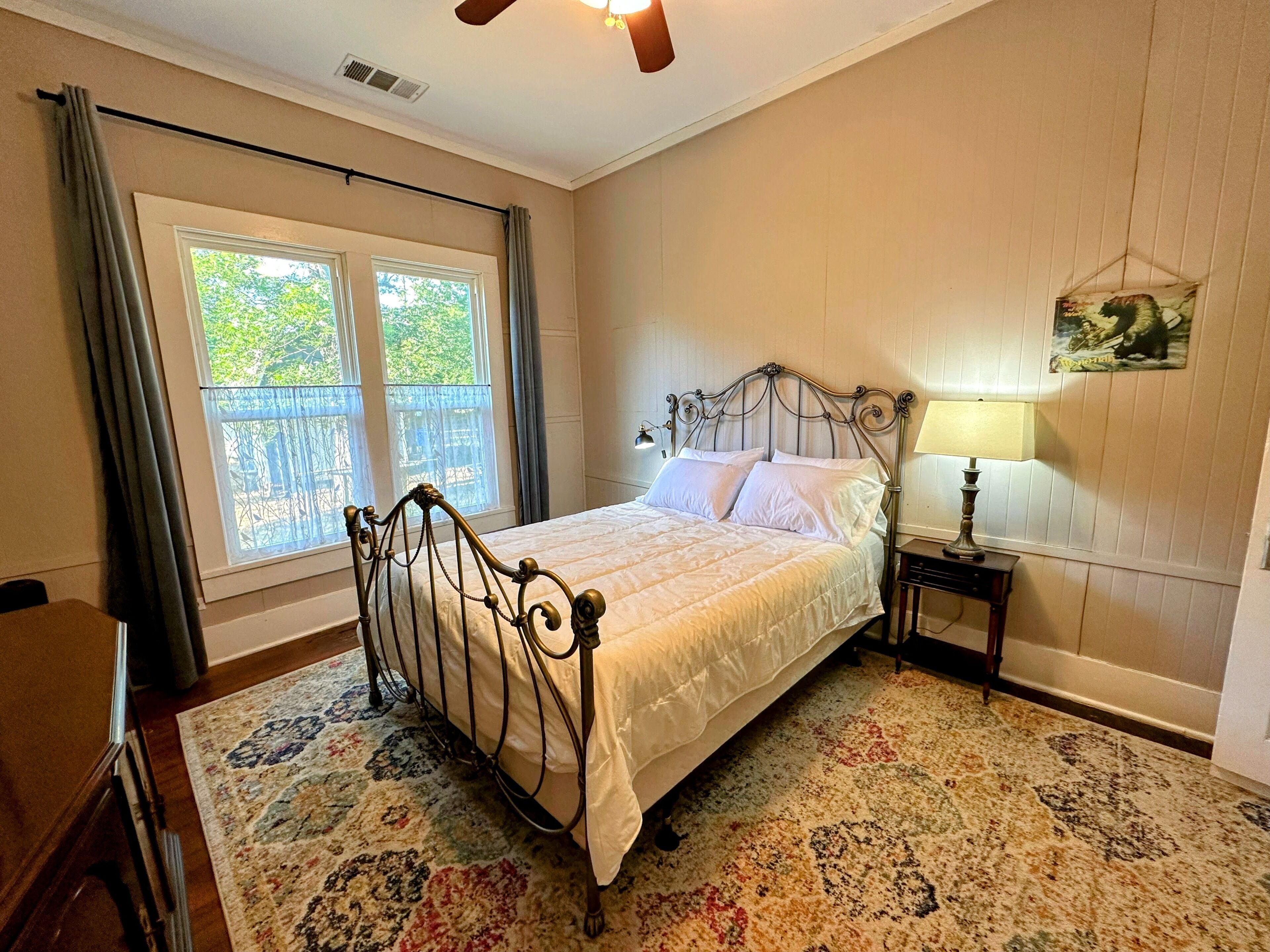 A bedroom with a bed, a nightstand, a lamp, a window with curtains, and a ceiling fan.