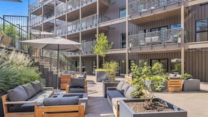 Un área de estar moderna al aire libre con muebles de madera y macetas, una escalera que conduce a un piso superior, y un edificio de varios pisos con balcones.