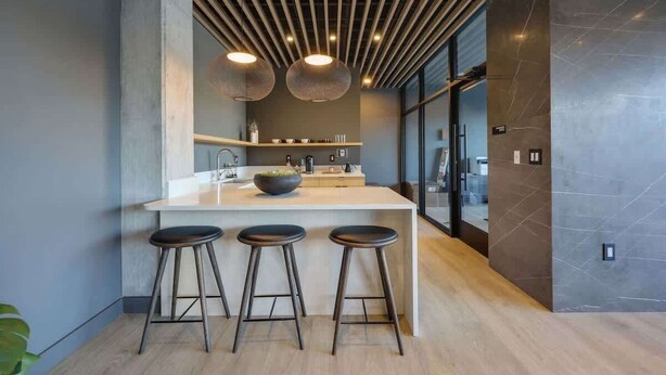 Una cocina moderna con una isla central, tres taburetes, iluminación colgante y un amplio ventanal.