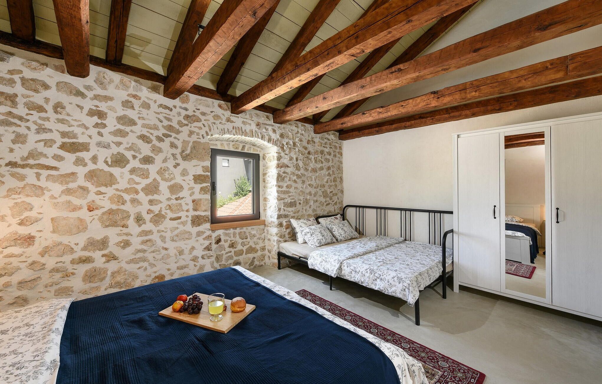 A bedroom with stone walls, a wooden ceiling, a bed with a blue bedspread, a bedside table with a tray of food, and a wardrobe.