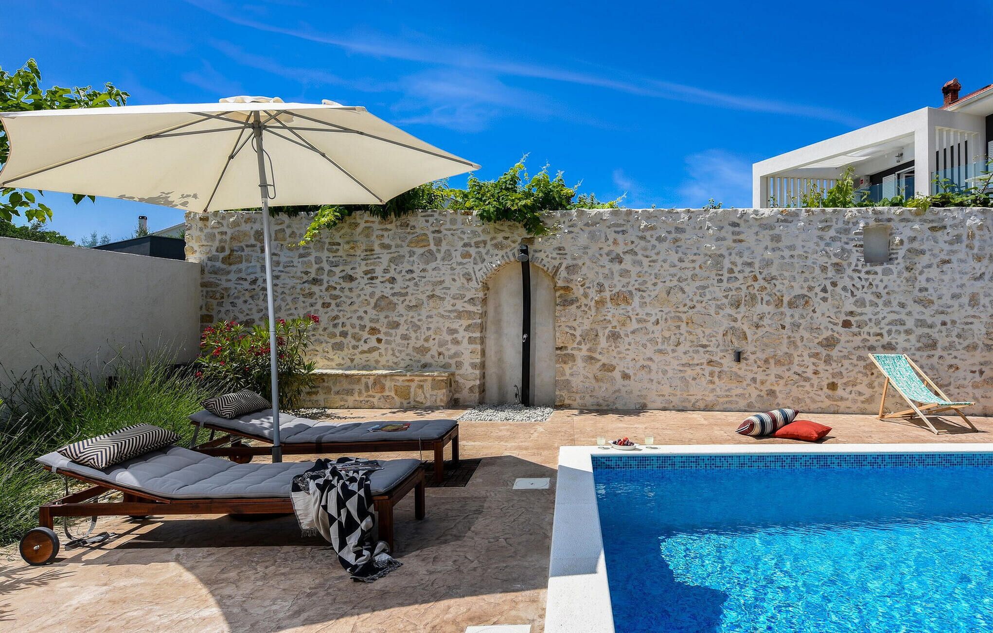 A pool area with lounge chairs, a covered umbrella, and a stone wall.