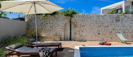A pool area with lounge chairs, a covered umbrella, and a stone wall.