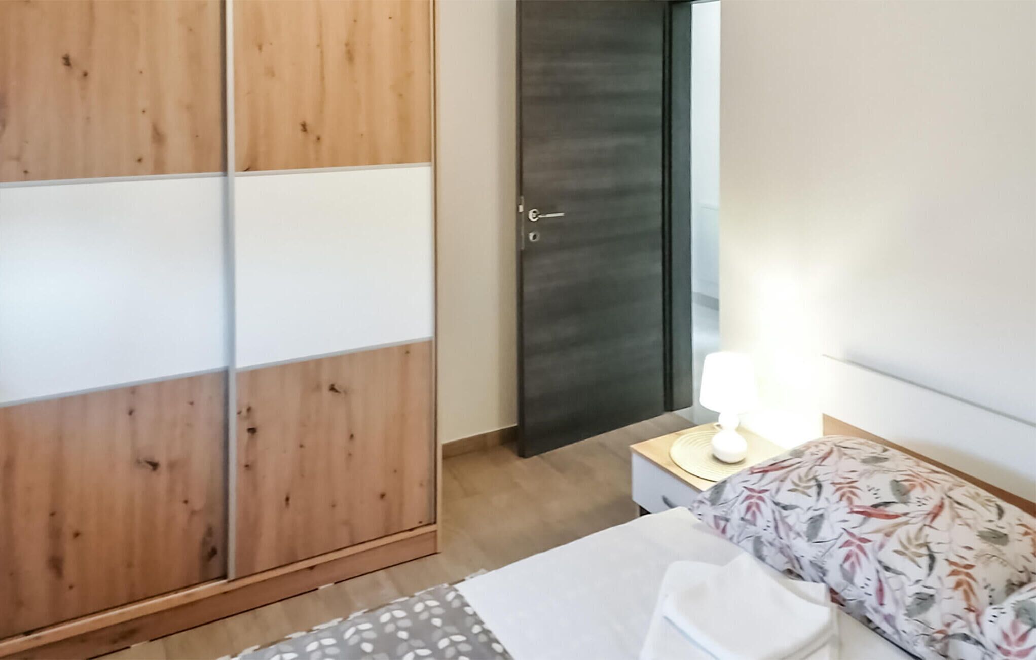 Un dormitorio con un armario de madera, una cama con colcha estampada, una mesita de noche con lámpara y una puerta oscura.