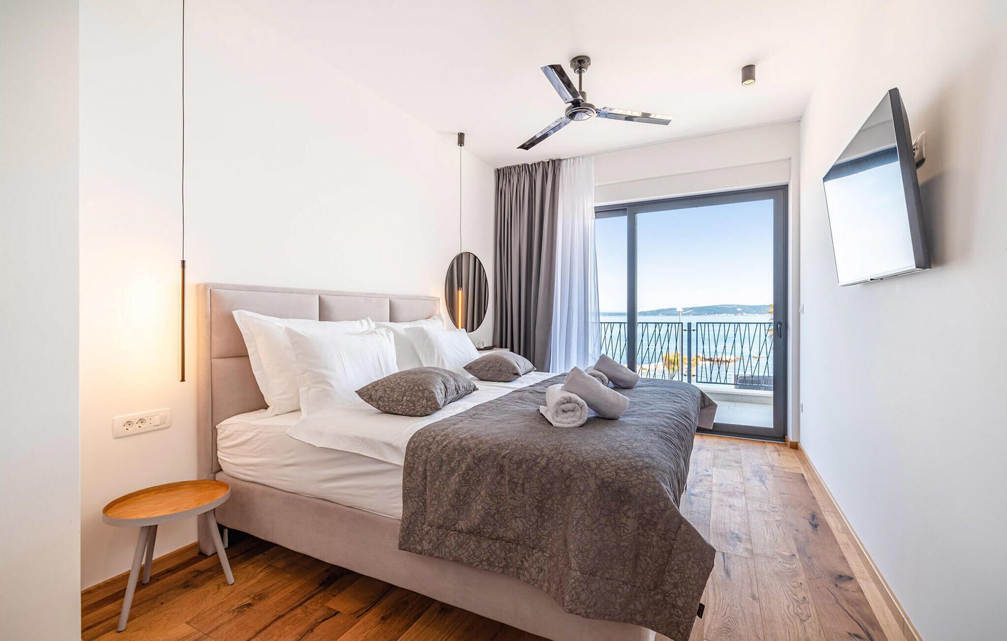 Een slaapkamer met een bed, nachtkastje, plafondventilator, televisie aan de muur en uitzicht op zee door een groot raam.