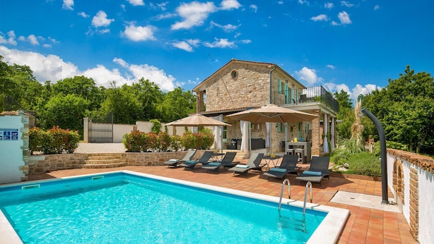 Une piscine avec des chaises longues et des parasols, un bâtiment en pierre doté d’un balcon, et un ciel d’un bleu limpide.