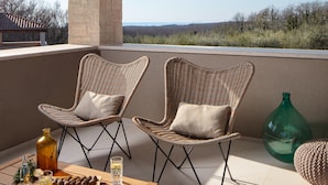 Un balcon avec deux fauteuils en osier, une table avec une bouteille, des verres et des amuse-gueules, et une vue sur le paysage.