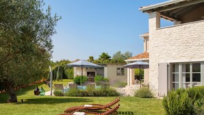Une maison avec une façade en pierre, une terrasse dotée de chaises longues et un jardin planté d’oliviers.