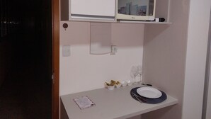 Piccola stanza con un armadietto bianco contenente una televisione e una mensola bianca sopra. C'è un tavolo con un piatto, una forchetta e dei bicchieri, e una piccola ciotola con degli snack.