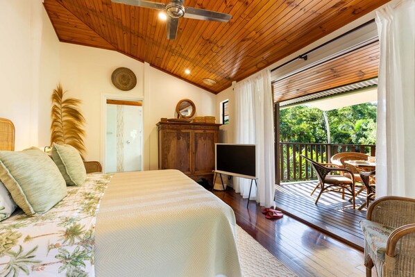 A bedroom with a bed, a wooden ceiling fan, a TV, and a balcony with wicker furniture.