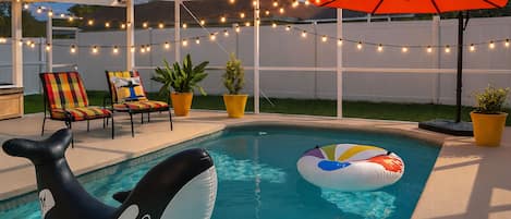 A backyard with a swimming pool, inflatable orca float, string lights, and outdoor furniture.