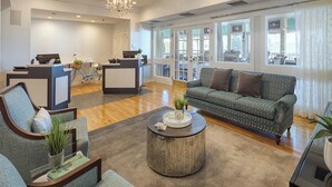 A spacious living area with a sofa, armchairs, and a coffee table. There is a reception desk with a bicycle and a computer. Large windows provide natural light and views of the outside. - 1 Bedroom Suite w/ Kitchenette, Sleeps 4, 10 min from Newport, at Voyager Inn! (Jamestown)