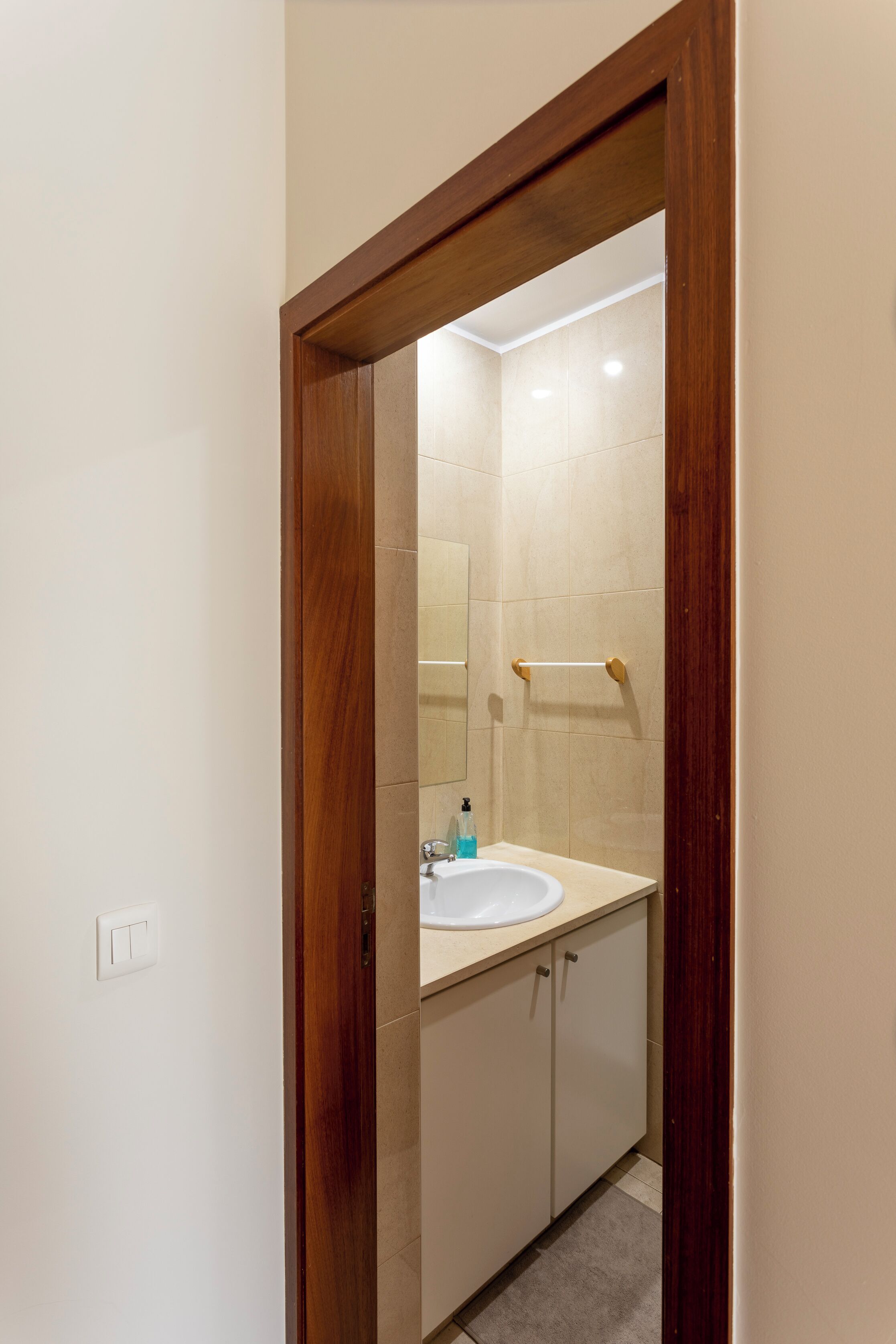 Un baño con puerta de madera, un lavamanos blanco, un espejo y baldosas color crema.