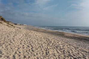 Ein sandiger Strand mit sanften Wellen, einem klaren Himmel und Gebäuden in der Ferne.