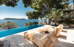 Zona junto a la piscina con vista al mar, mobiliario exterior y una palmera.