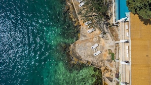 Luftaufnahme eines Küstenhotels mit Pool, Liegestühlen und felsiger Küstenlinie.