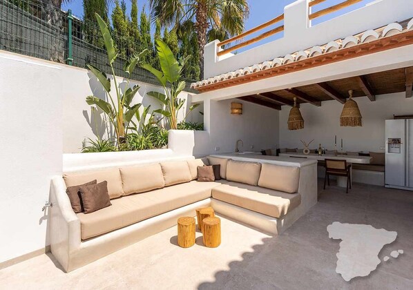 An outdoor living area with a beige sectional sofa, wooden stools, and a kitchen area with hanging lights.