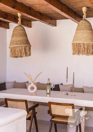 A dining area with a white table, two wicker pendant lights, a bottle of champagne, and candles.