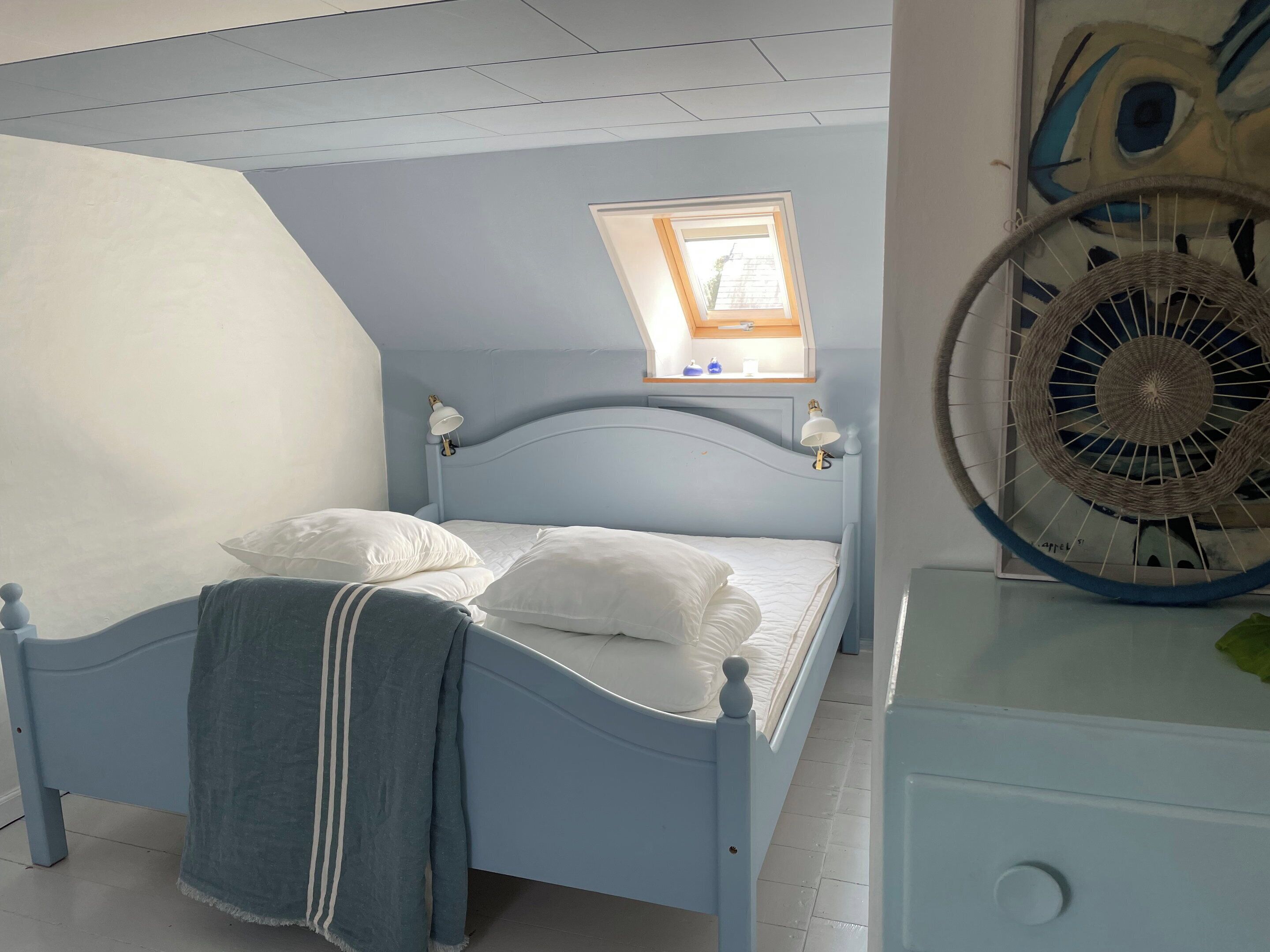 Une chambre à coucher avec un lit, une fenêtre, un ventilateur fixé au mur et une petite étagère avec quelques objets.