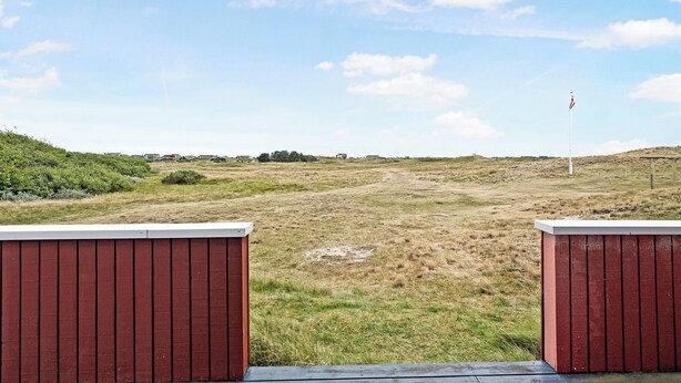 A view from a covered area with a red wooden structure, a grassy field, a flag, and distant buildings.