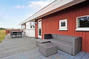 Un espacio exterior cubierto con una terraza de madera, un sofá de mimbre y una mesa de comedor.