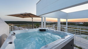Een jacuzzi op een balkon met uitzicht op een zonsondergang, witte leuningen en lounge stoelen.