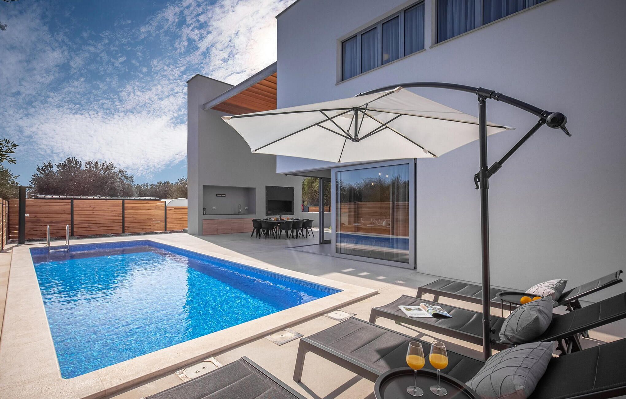 Ett modernt utomhusområde med pool, ett inbyggt kök och en matplats, omgivet av ett trästaket.
