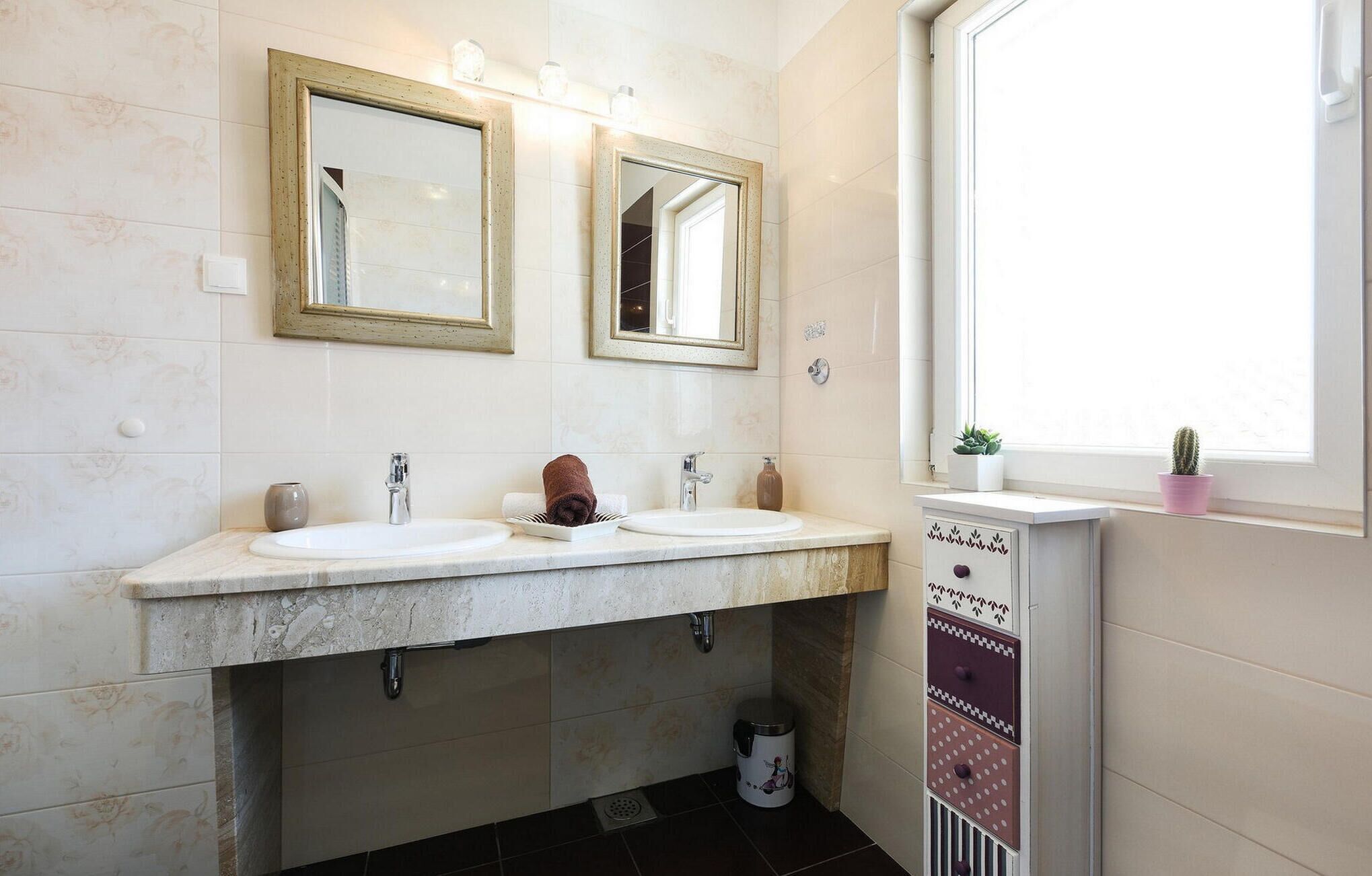 A bathroom with two sinks, a window with plants, and a trash can.