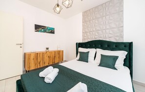 A neatly made bed with a green headboard, white bedding, and matching pillows. A wooden nightstand with a plant and a lamp is beside the bed. - Beach front home in Razanj (Razanj)