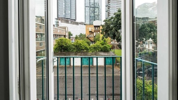 Une vue depuis une fenêtre donnant sur un parking délimité par une clôture verte, un bâtiment à la façade incurvée et, au loin, un grand immeuble d’habitation.