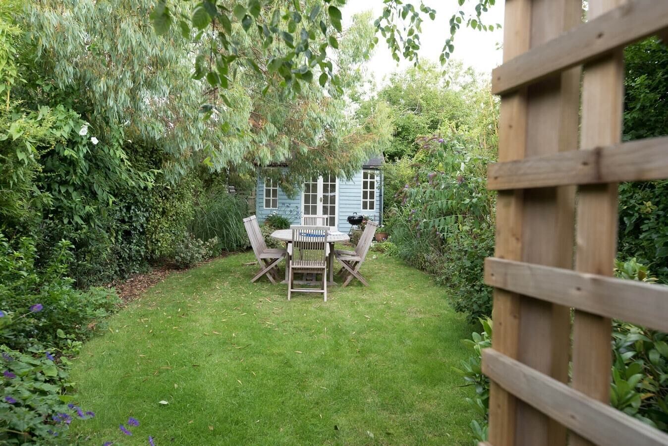 Een tuin met een houten tafel en stoelen, omringd door weelderige begroeiing en een blauwe schuur.