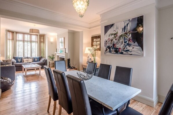A spacious living room with a dining area, featuring a large painting, a fireplace, and a chandelier.