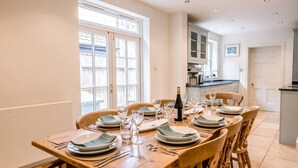 A dining area with a wooden table set for four, large windows, and a kitchen in the background. - 6 The Terrace, Walberswick (Walberswick)