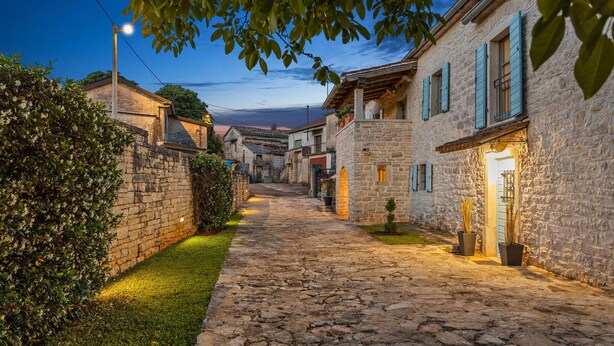 Una estrecha calle adoquinada bordeada por edificios de piedra con contraventanas, que conduce a una puerta.