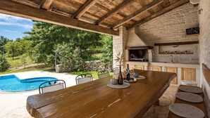 Cocina al aire libre con pared de piedra, piscina y mesa de madera.