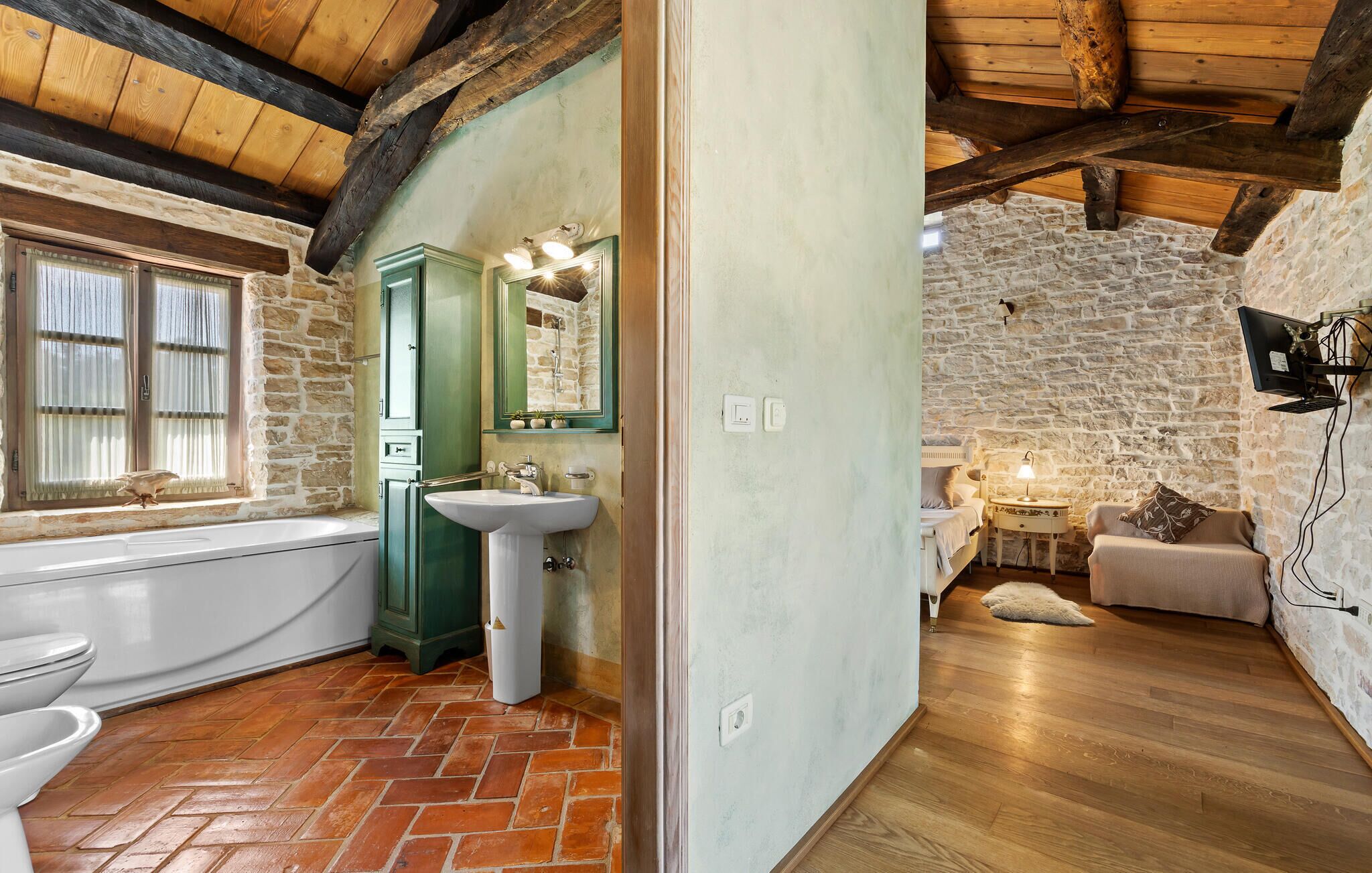 A rustic bathroom with a stone wall, wooden beams, a green cabinet, a sink, and a bathtub.