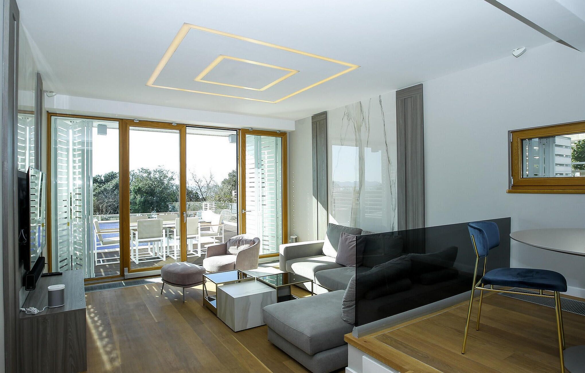 A modern living room with a sofa, a coffee table, and a TV. There is a balcony with outdoor furniture.