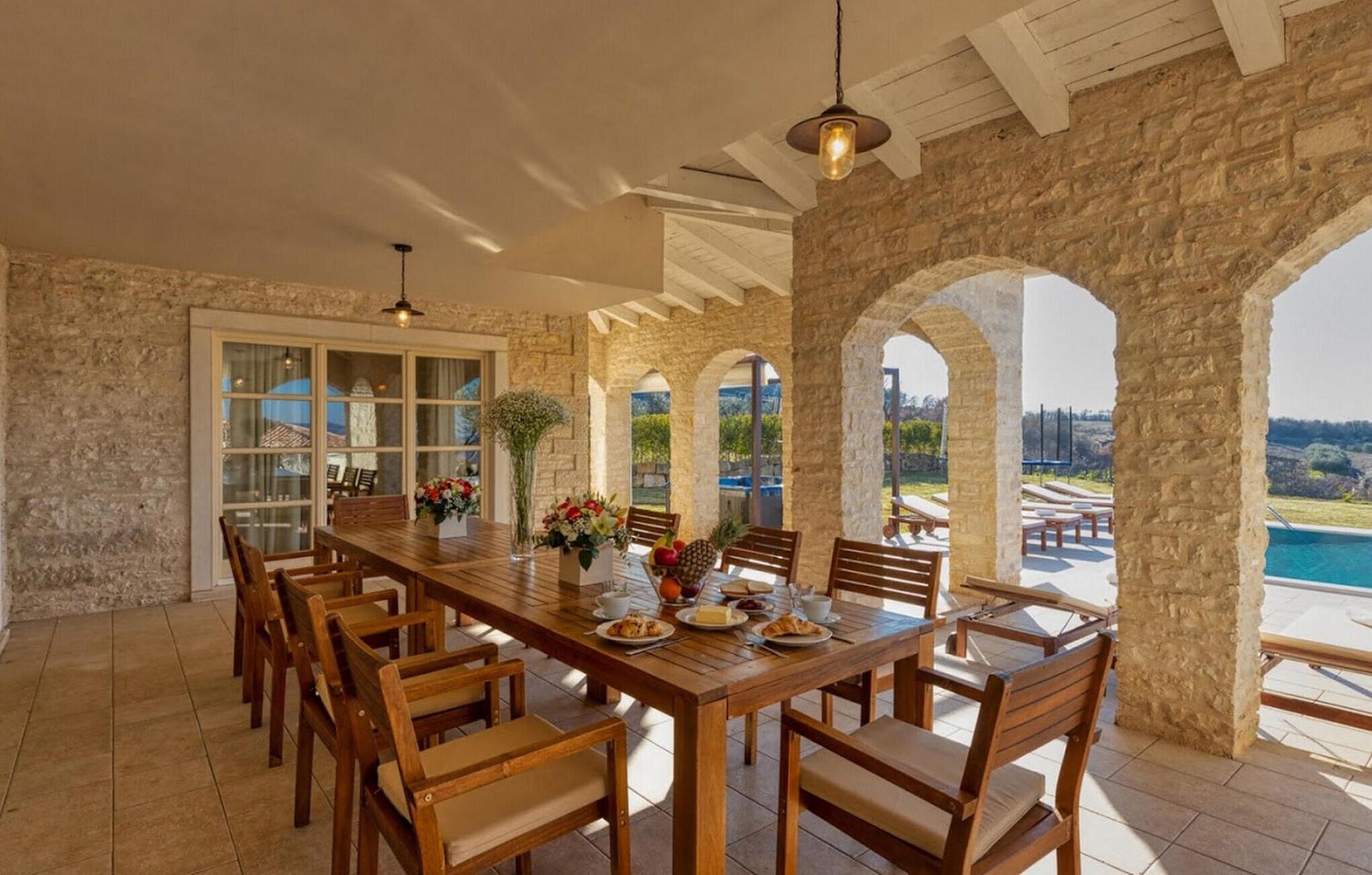 A spacious dining area with a long wooden table and chairs, stone walls, and large windows offering a view of a pool and landscape.