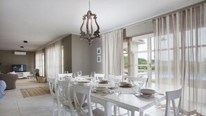 Een eetruimte met een witte tafel gedekt voor vier personen, omringd door witte stoelen, een kroonluchter en grote ramen met lichte gordijnen.