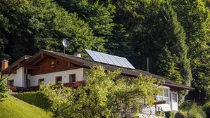 Una casa de fusta amb panells solars a la teulada, envoltada de vegetació abundant i arbres.