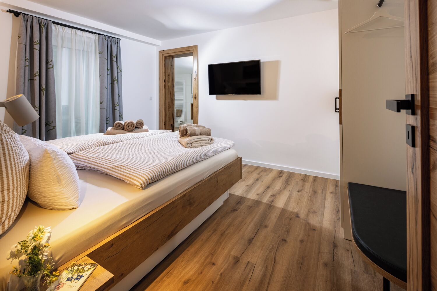 A modern bedroom with a bed, wooden floor, a mounted TV, and a bedside table with a lamp.