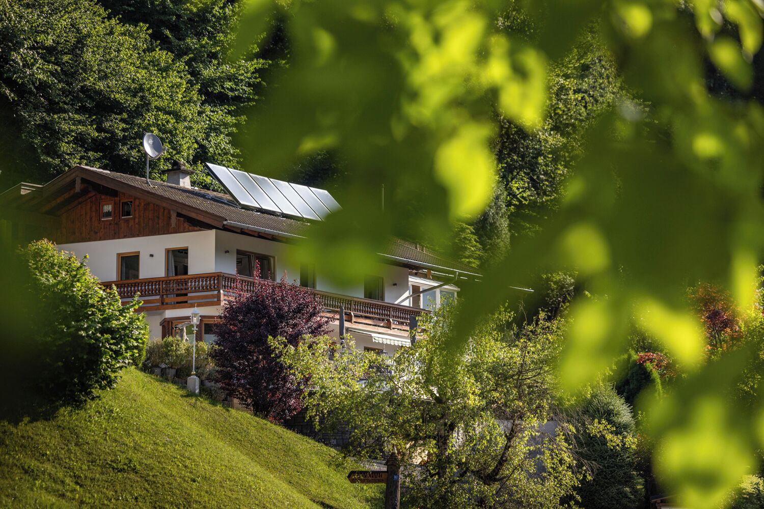 A two-story house with a balcony, surrounded by lush greenery and a sloped roof with solar panels.