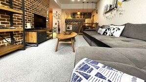 Una sala de estar moderna con pared de ladrillo, un sofá gris, una mesa de centro de madera y un televisor sobre una estantería.