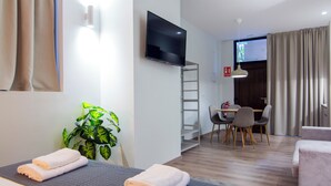 Une chambre d’hôtel comprenant un lit, un téléviseur fixé au mur, un bureau avec des chaises, une plante en pot et un coin repas agrémenté de rideaux.