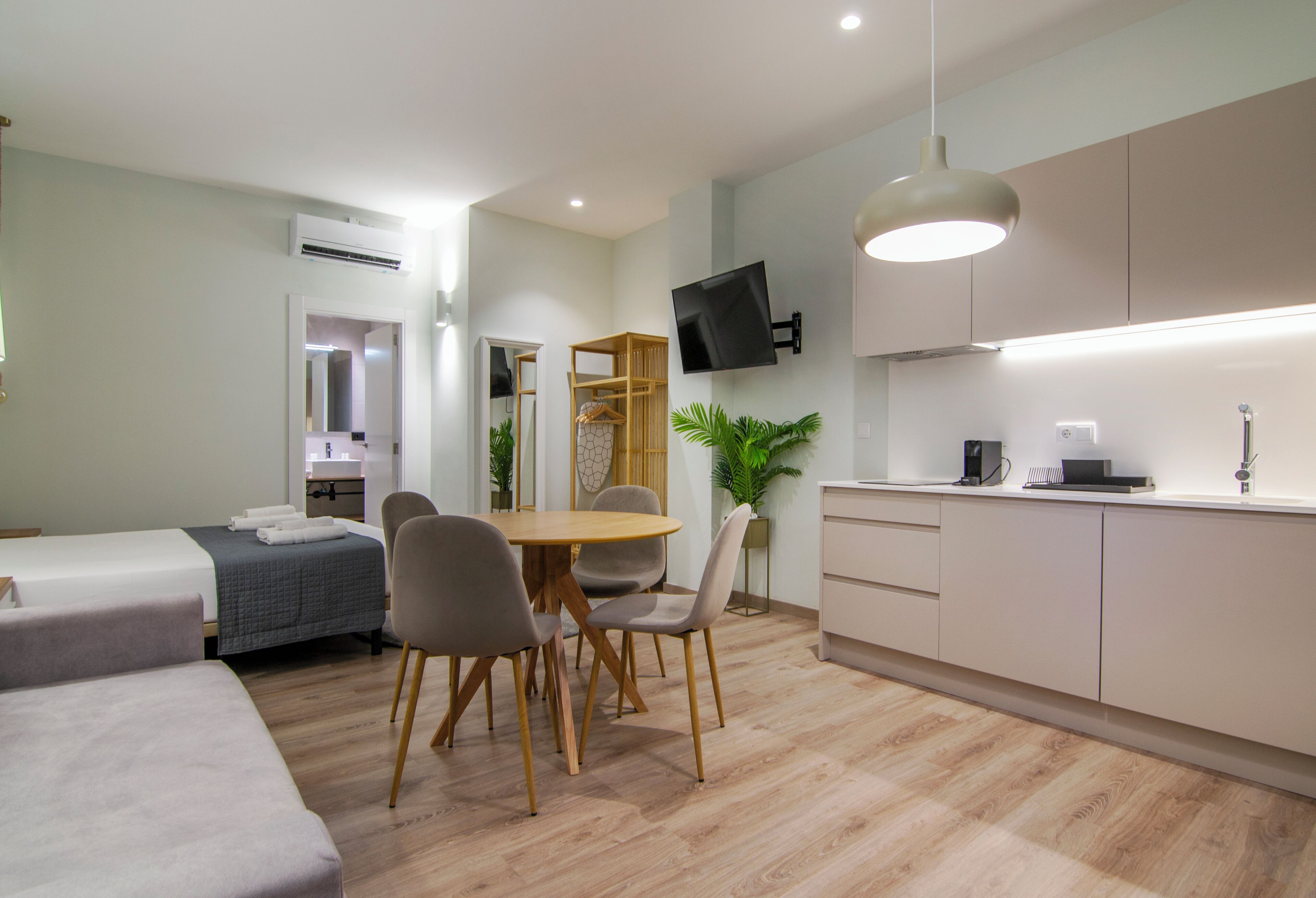 A modern hotel room with a bed, a dining area with chairs, a small kitchen with a sink, and a TV mounted on the wall.