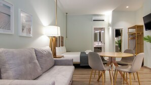 Una habitación de hotel moderna con un sofá gris, una mesa de comedor de madera con cuatro sillas, un televisor de pantalla plana en la pared y una pequeña kitchenette.