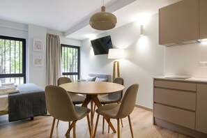 A modern hotel room with a round dining table, four chairs, a bed with a gray quilt, a TV mounted on the wall, and a kitchen area with cabinets and a lamp. - VLC HOST - Reino 7 (Valencia)