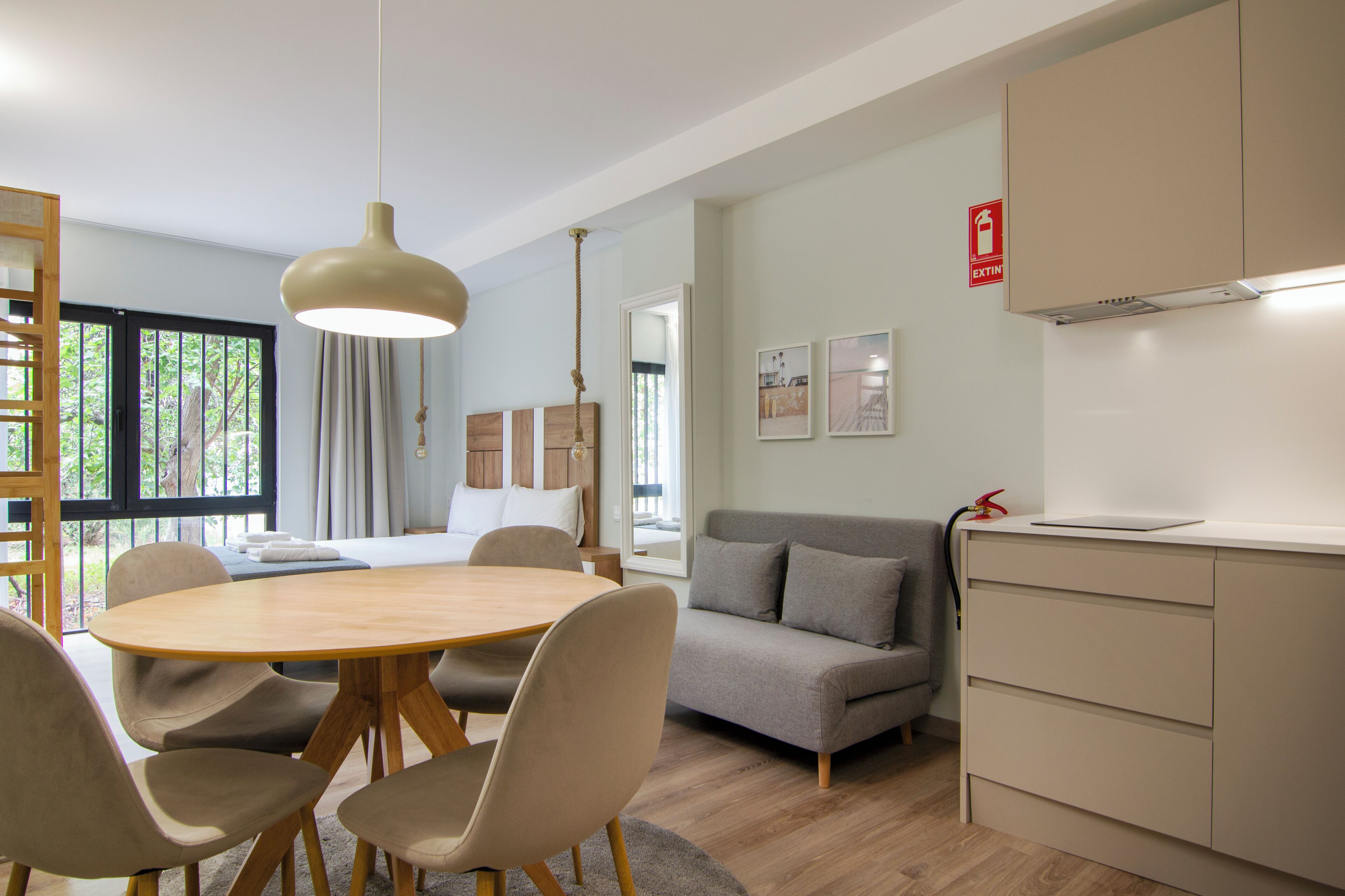 Une pièce moderne avec une table de salle à manger ronde en bois, des chaises beiges, un canapé gris et une kitchenette dotée d’un plan de travail blanc.