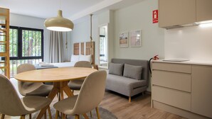 Una habitación moderna con una mesa de comedor de madera redonda, sillas color beige, un sofá gris y una kitchenette con encimera blanca.